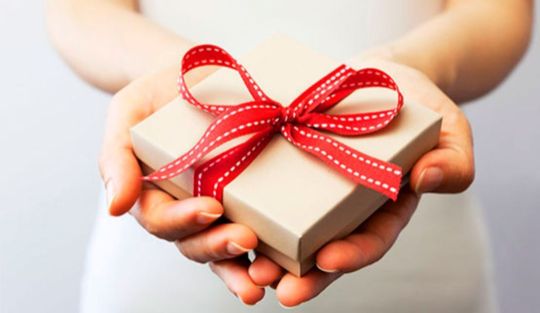 Девушка, на уровне пояса, нежно держит в ладошках двух рук подарок в подарочной упаковке. Ладошки и подарок сложены в виде сердечка 